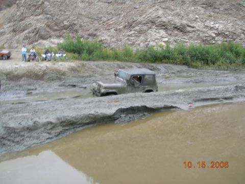 1985 Ford Bronco - A Jeep Wrangler navigates a muddy trail, while other vehicles and spectators watch from the embankment.
