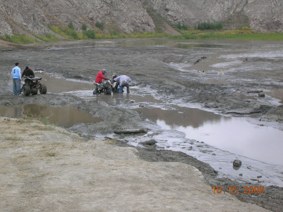 Two ATVs are in a muddy, wet area with people around them. Another ATV is parked nearby with two people standing next to it