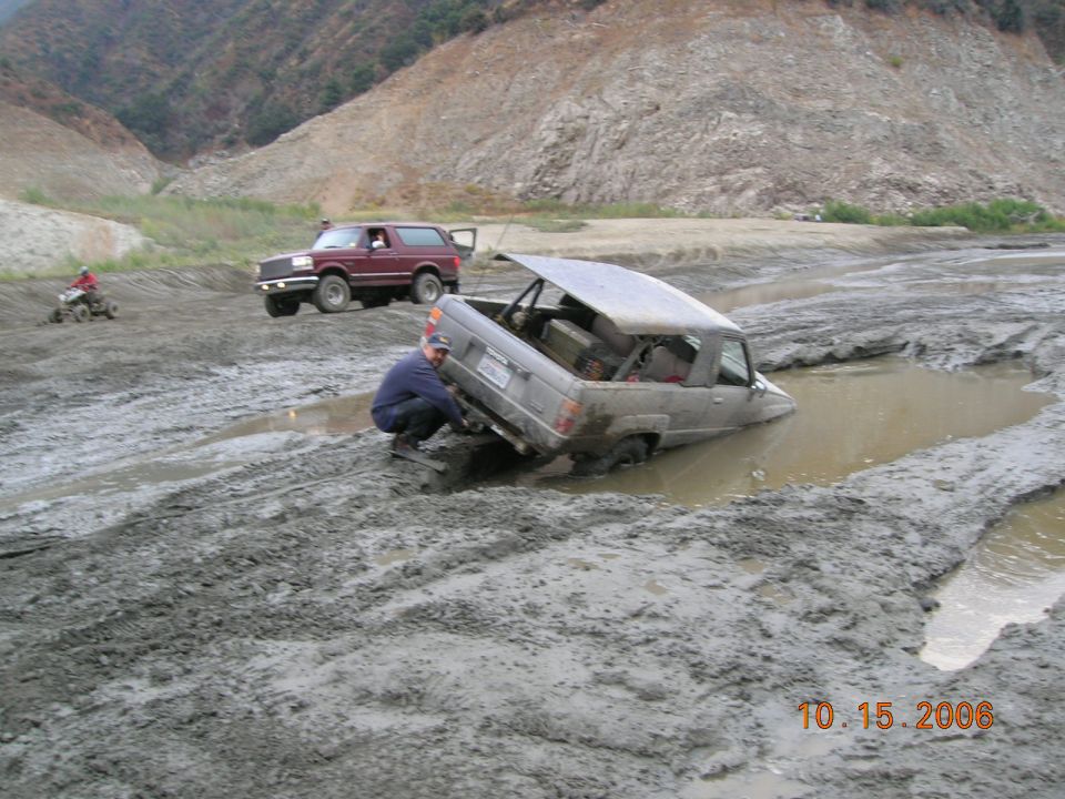 Two off-road vehicles stuck in mud, one partially submerged. A person is attempting to free the submerged vehicle
