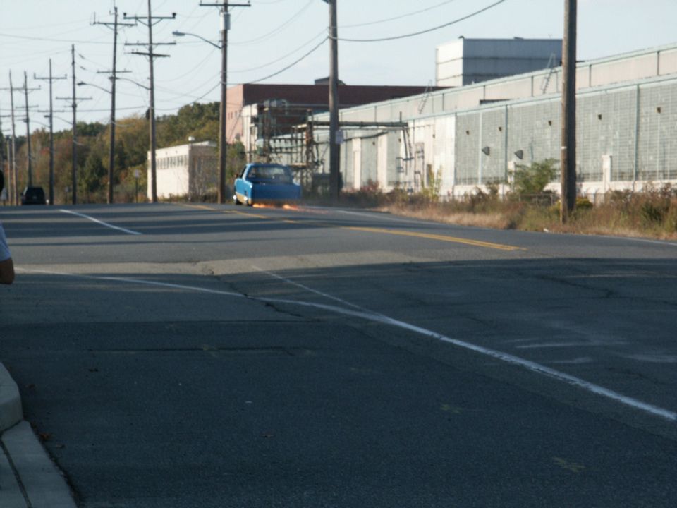 Blue car with flames erupting from the rear bumper, driving down a road