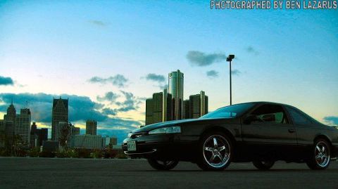 1997 Ford Thunderbird - A black 1997 Ford Thunderbird coupe with custom chrome wheels is parked with the Detroit skyline in the background.