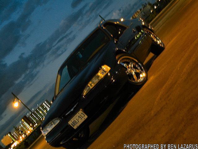 Black sedan with custom rims parked on a street at dusk, city lights in background