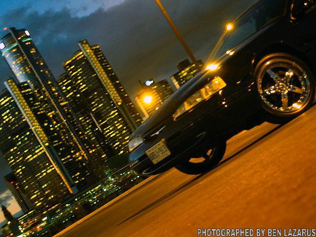 Black 1997 Ford Thunderbird with aftermarket wheels, seen from a 3/4 front angle at night with city lights in the background
