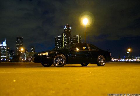 1997 Ford Thunderbird - A black coupe is parked on a yellow surface with a city skyline illuminated at night.
