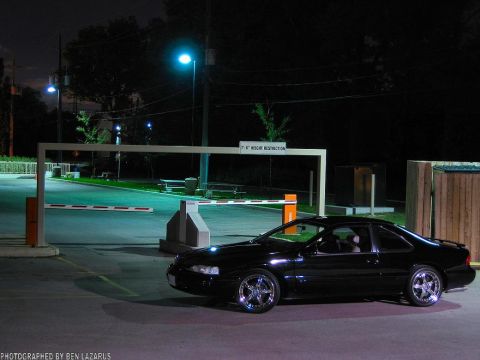 1997 Ford Thunderbird - A black coupe with custom chrome wheels is seen parked at night near a height restriction gate.