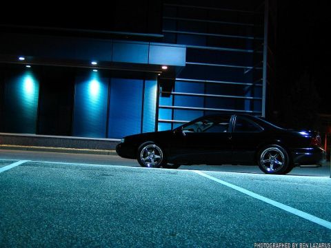 1997 Ford Thunderbird - A black coupe is parked in front of a modern building at night, illuminated by blue lights.