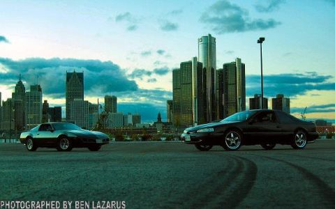 1997 Ford Thunderbird - Two coupes are parked with the Detroit skyline in the background under a cloudy sky.
