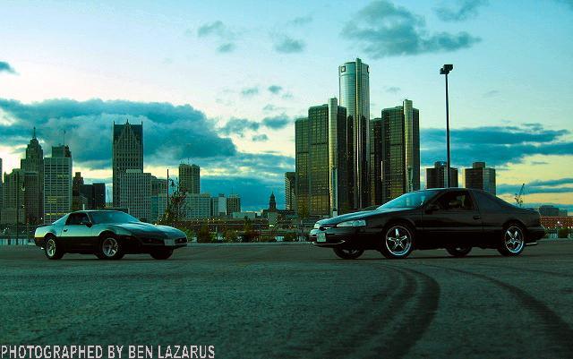 Black coupe and a lighter coupe parked with Detroit skyline behind them