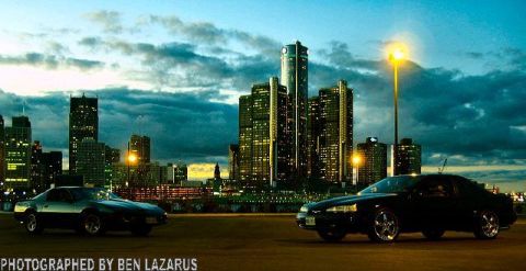 1997 Ford Thunderbird - Two cars are parked in front of a city skyline as the sky darkens.