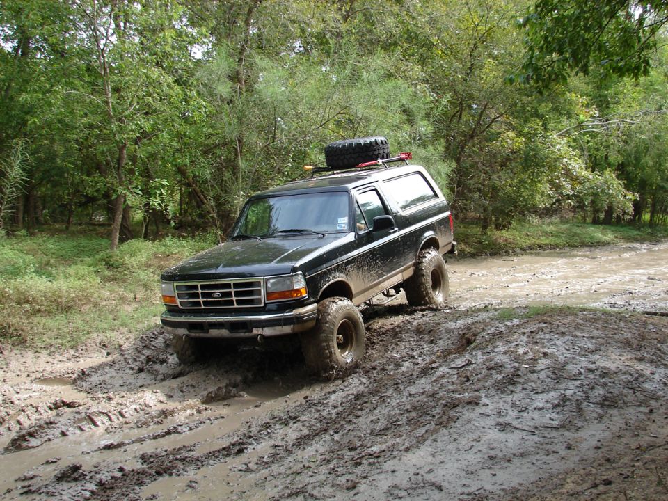 Black 1996 Ford Bronco driving through a muddy trail with a spare tire on the roof rack