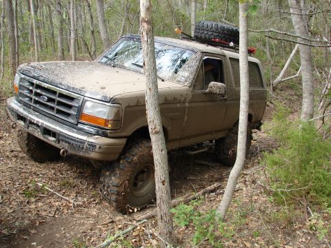 1996 Ford Bronco - A 1996 Ford Bronco navigates a muddy, wooded path with a spare tire mounted on its roof rack.