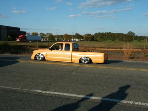 2001 Mercury Grand Marquis - An orange pickup truck with a custom lowered suspension drives down a two-lane road under a clear blue sky.