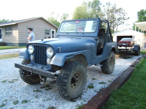 1980 Jeep CJ5 - Blue Jeep CJ5 is parked on gravel, towing a trailer with a red car. A man stands near a house in the background.
