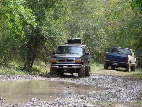 1996 Ford Bronco - A dark 1996 Ford Bronco navigates a muddy trail with a spare tire mounted on its roof.