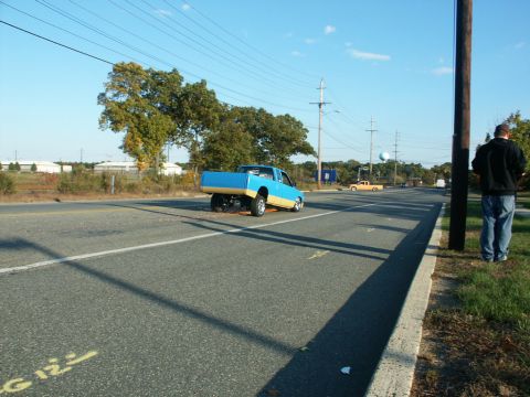 2001 Mercury Grand Marquis - A blue and tan pickup truck is seen on the road with its rear end lifted, while another yellow truck is visible in the distance.