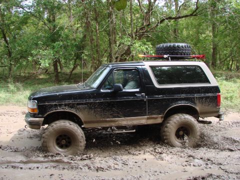 1996 Ford Bronco - A black Ford Bronco navigates a muddy trail with large tires, a spare tire mounted on its roof rack.