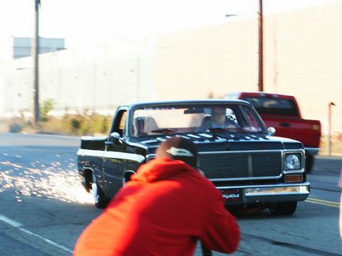 2001 Mercury Grand Marquis - A black pickup truck with sparks flying from its rear wheel is captured in motion, with another red truck visible in the background.