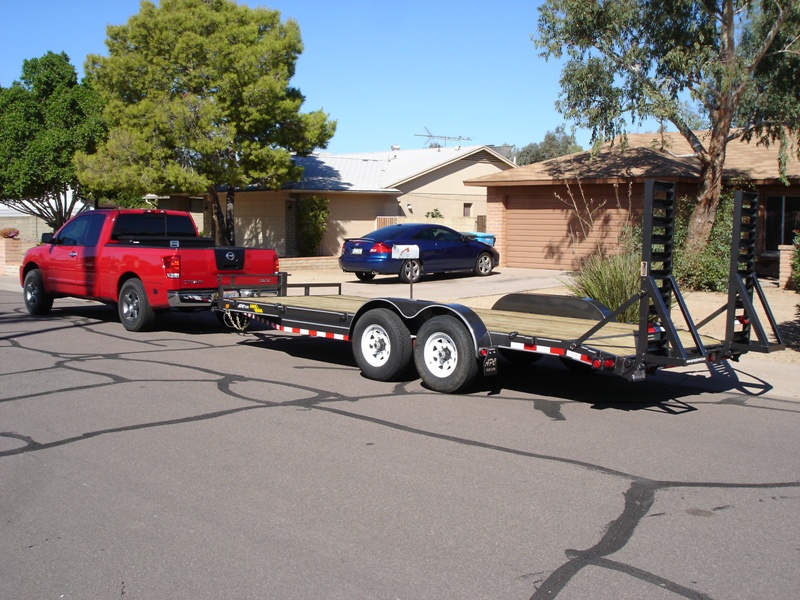 Red Nissan pickup towing an empty black tandem-axle car trailer on a street