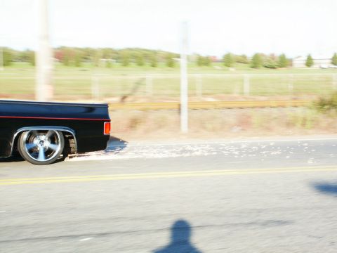 2001 Mercury Grand Marquis - A black pickup truck with custom wheels drives down a road, with a blurred background of trees and a fence.
