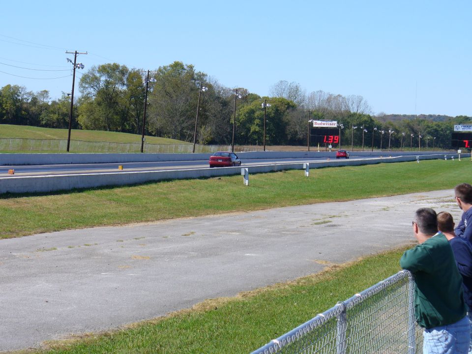Red car and a darker car race on a drag strip with a digital timer showing 1:39