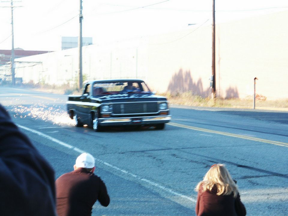 Black pickup truck doing a burnout on a road, sparks flying from rear wheel. Two people watching
