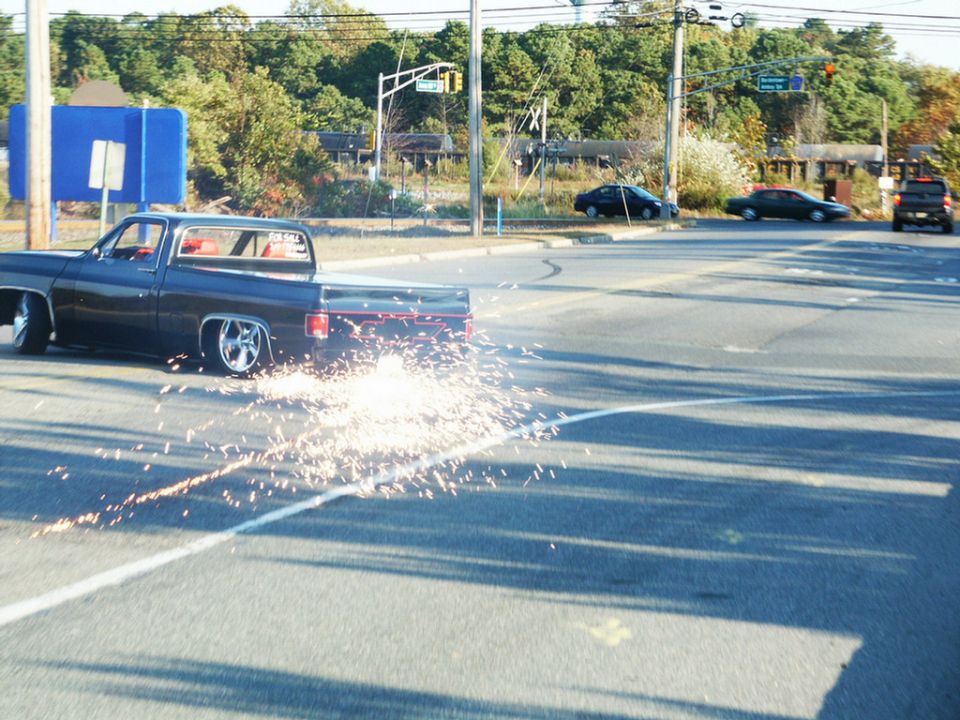 Lowered dark blue pickup truck doing a burnout, sparks flying from rear tires. Other cars visible in background