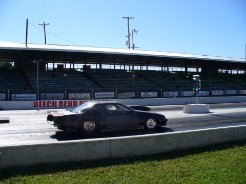 1991 Ford Probe - A dark coupe drag car races down the track at Beech Bend Raceway, with grandstands in the background.
