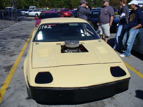 1991 Ford Probe - A modified yellow car with an exposed engine sits on a track, with other vehicles and people in the background.