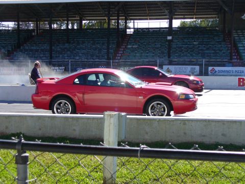 1991 Ford Probe - Two red Ford Mustangs race down a drag strip with grandstands in the background.
