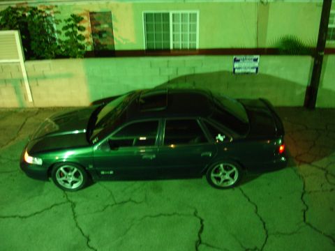 1993 Ford Taurus - A dark green sedan is parked on cracked pavement at night, with a building and some greenery behind it.