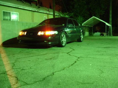 1993 Ford Taurus - A dark sedan is parked on cracked asphalt at night, with its headlights illuminated.