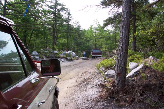 View from inside a vehicle looking down a dirt road with another SUV ahead. Trees and rocks line the path