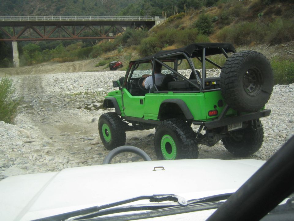green Jeep with big tires and roll cage driving on a rocky riverbed, another red vehicle in background