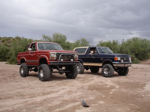 1984 Ford Bronco - Two modified Ford Broncos sit side-by-side in a dusty, outdoor setting under a cloudy sky.