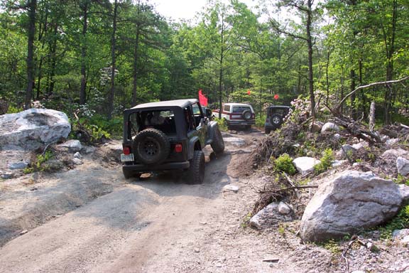 Jeep Wrangler on a dirt trail with other SUVs in the background, surrounded by trees and rocks