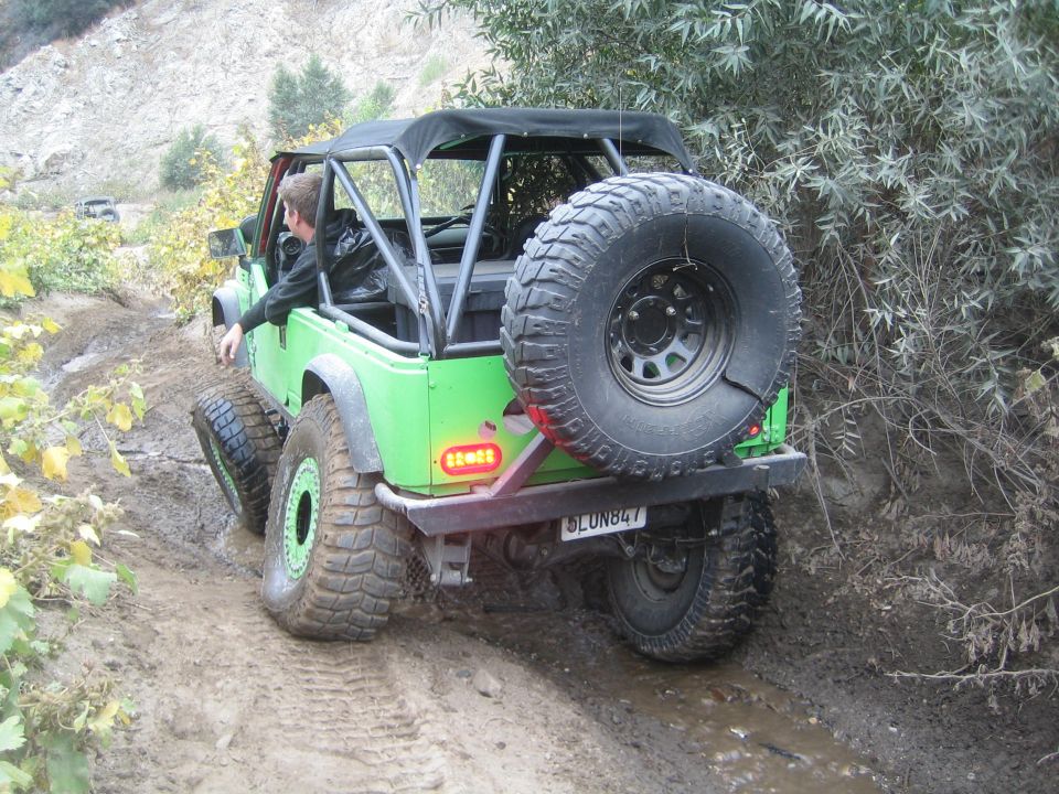 Green Jeep Wrangler modified for off-roading, driving on a muddy trail. Spare tire mounted on the rear. Another vehicle is visible in the background