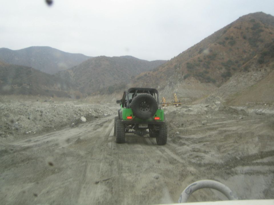 Green Jeep Wrangler driving on a dirt road in a mountainous, barren area. Another vehicle is partially visible in the foreground