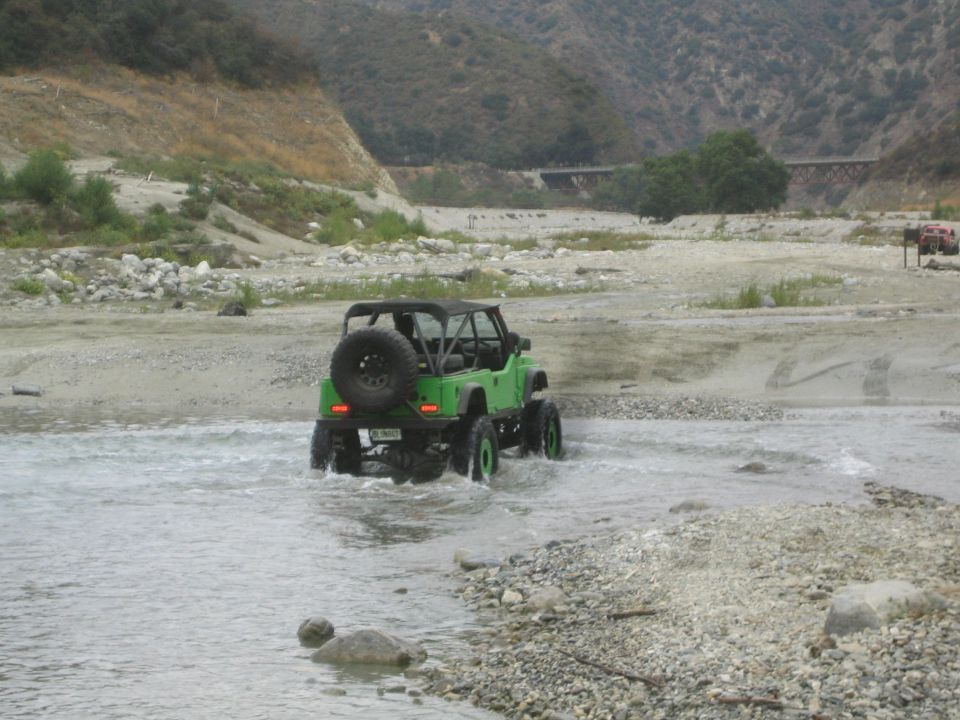 Green off-road vehicle with spare tire on back crossing a shallow riverbed. Another red vehicle is visible in the distance