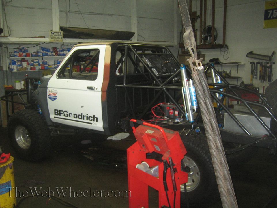 Off-road race truck with roll cage, BFGoodrich tires, and Lucas Oil decals. It is in a workshop with tools and parts visible