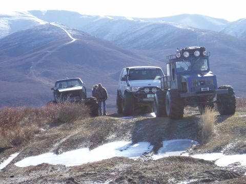 2000 Ford E350 - Three modified off-road vehicles are parked on a muddy and snowy hillside with mountains in the background.