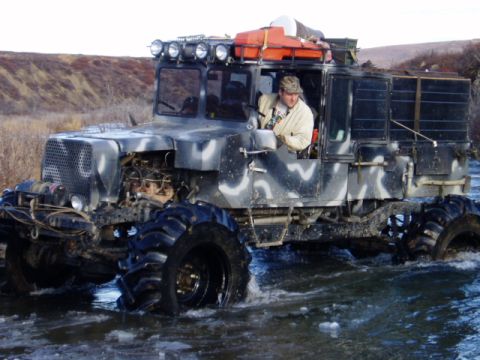 2000 Ford E350 - An off-road vehicle with a camouflage paint scheme navigates through shallow water, its large tires churning the surface.