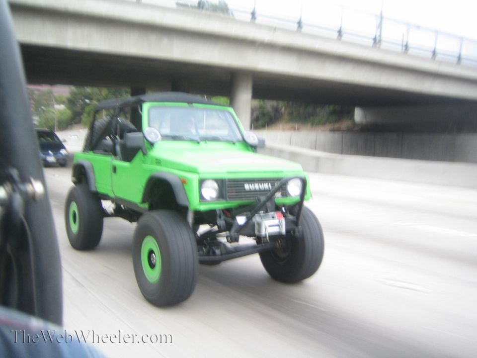Green Suzuki Samurai with big tires driving on highway. Another car is visible in the background