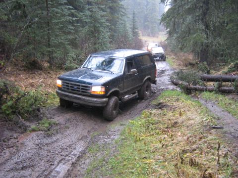 1995 Ford Bronco - A 1995 Ford Bronco drives through mud on a forest road, with another vehicle in tow.
