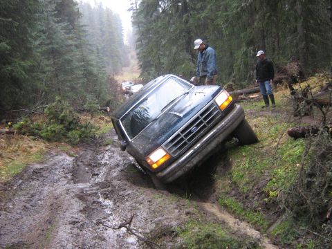 1995 Ford Bronco - A dark blue Ford Bronco is stuck in the mud on a forest road, with two people observing.
