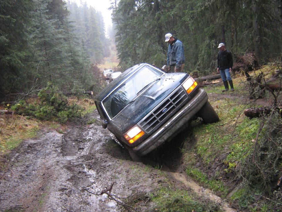 Dark green Ford Bronco leaning into a deep muddy rut on a wet forest trail