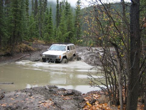 1995 Ford Bronco - A Jeep Cherokee XJ navigates a deep mud puddle on an off-road trail surrounded by dense forest.