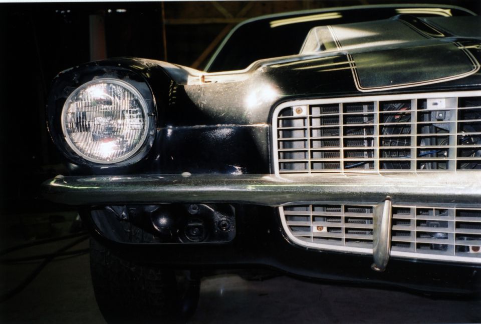Close-up of dark green Camaro front end with round headlight, chrome bumper and grille in a garage