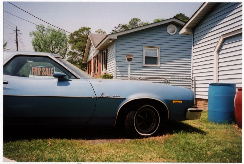 Light blue Ford Ranchero side view with FOR SALE sign in window, parked on grass by a blue house