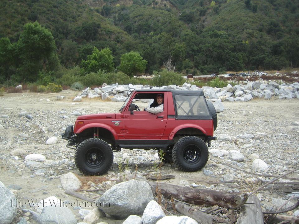 Red 1987 Suzuki Samurai with lifted suspension and large tires parked on a rocky, dry riverbed with a wooded hillside in the background