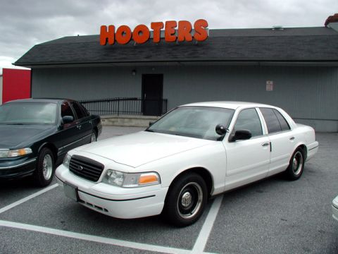 1998 Ford Crown Victoria - A white 1998 Ford Crown Victoria is parked in front of a Hooters restaurant, with another dark sedan to its left.